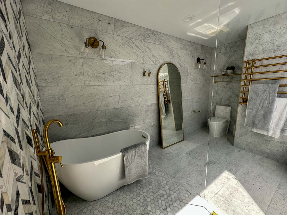 A minimalist bathroom with a toilet, a full body mirror, tiled floor and backsplash, freestanding bathtub, and a towel bar