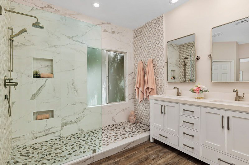 A bathroom with wide shower area that has a glass partition, tiled backsplash, a vanity area with storage cabinet, sinks, and mirrors