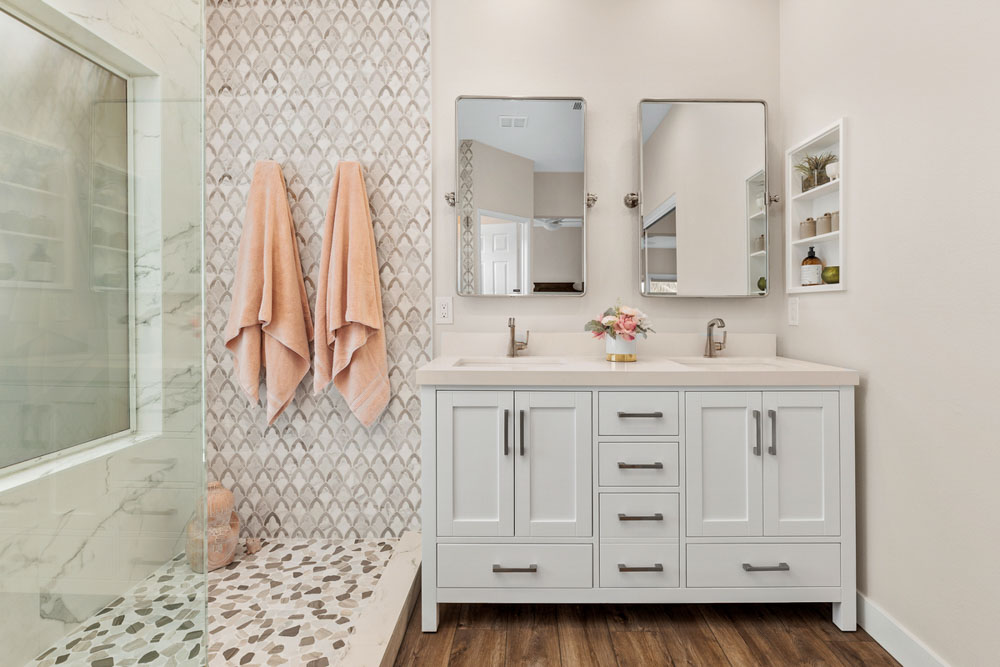 A remodeled bathroom with shower area that has a glass partition, tiled backsplash, and floor, and a storage cabinet with countertop and sink, two mirrors, and a recessed shelf for bath necessities
