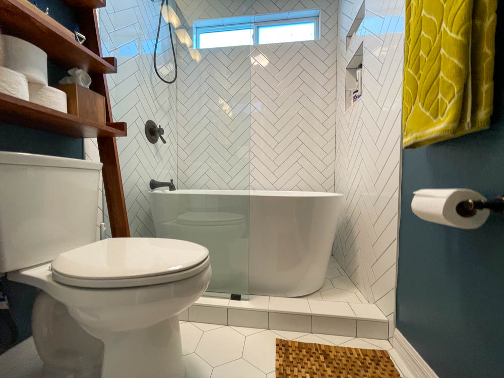 A renovated bathroom with tiled backsplash, and floor, a freestanding bathtub, and wooden shelf with bathroom necessities.