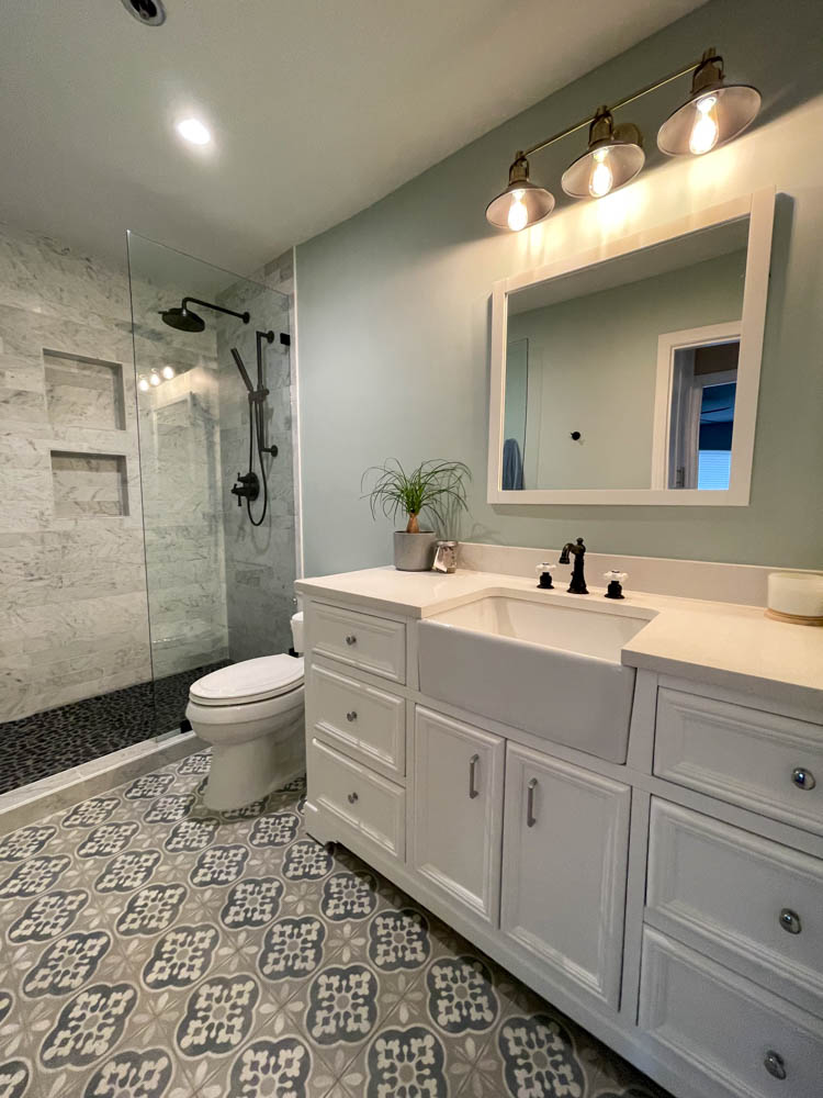 A remodeled bathroom with white storage cabinets, patterned tile flooring, and a tile backsplash, and a glass partitioned shower area.