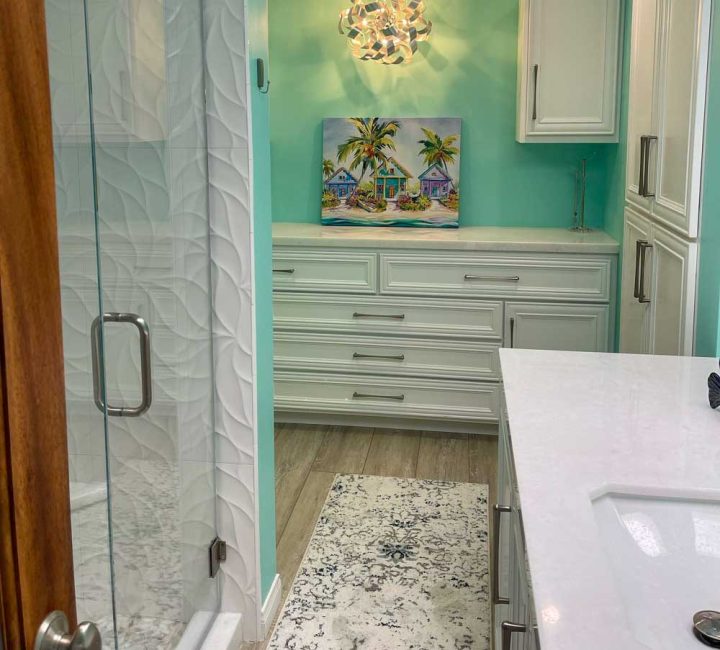 A relaxing bathroom with mint green painted wall, a shower area enclosed in glass, a sink on a ceramic countertop of a storage cabinet, white drawers and cabinets with tropic painting on top.
