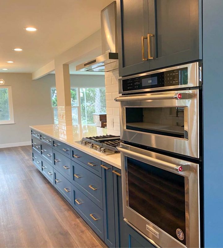 A remodeled kitchen with blue cabinets, ovens, a built-in cooktop with steel exhaust hood and a ceramic countertop